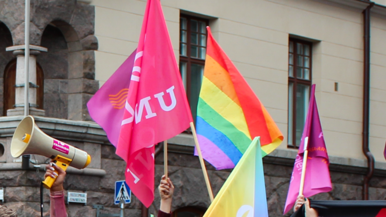 Naisten kädet pitelevät naisjärjestöjen lippuja sekä Pride-lippua ja megafonia.